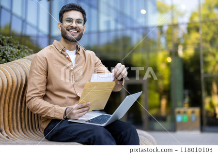Young adult wearing glasses holds envelope with smile, seated on outdoor bench with laptop open. Bright urban setting conveys success, positivity, work-life balance. 118010103