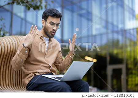 Young man sitting outdoors, displaying frustration with laptop. Occurs in urban environment showcasing unexpected technical problem. Concept of technology mishap, communication, and workplace stress 118010105
