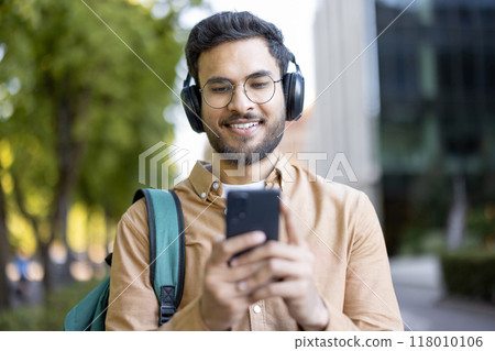 Young man with headphones using smartphone outdoors. Enjoying music, wearing casual attire with backpack. Displaying relaxation, enjoying day. Captures lifestyle, modern technology, student life. 118010106