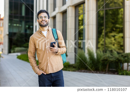 Confident young Indian student holding smartphone, smiling, and standing outdoors on a sunny university campus. Concept of education, youth, and technology. 118010107