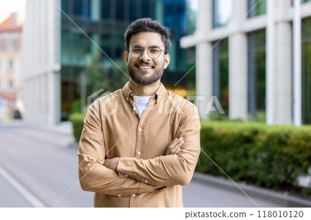 Young man with glasses standing confidently outside modern office building. Smiling and crossing arms, conveying confidence and friendliness. Background features urban environment 118010120