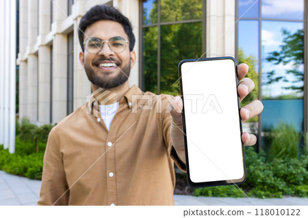 Smiling man with glasses holding blank phone screen outdoors near modern office building. Concept of technology, communication, and business. Ideal for promotional content or mockup design. Smiling man with glasses holding blank phone screen outdoors near modern office building. Concept of technology, communication, and business. Ideal for promotional content or mockup design. 118010122