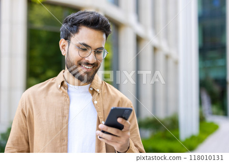 Man wearing glasses and casual clothing smiling while using smartphone outdoors near modern building. Person enjoying communication and technology in outdoor setting on sunny day 118010131