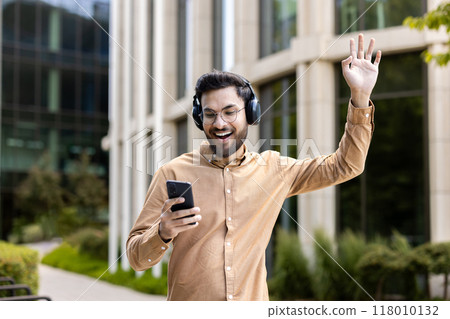 Smiling man wearing headphones uses smartphone enjoying music outdoors. Standing with hand raised in excitement, expressing happiness and enjoyment in sunny urban setting. Smiling man wearing headphones uses smartphone enjoying music outdoors. Standing with hand raised in excitement, expressing happiness and enjoyment in sunny urban setting. 118010132