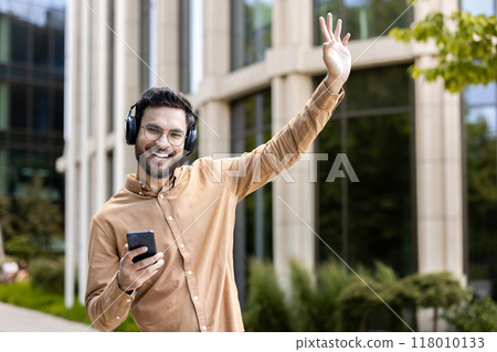 Cheerful man wearing headphones smiling while holding phone and waving outside modern building. Person enjoying music and technology in casual setting, conveying happiness and connection in outdoor 118010133