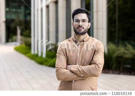 Confident young man with glasses in casual shirt standing with arms crossed outside modern office building. Represents professionalism, confidence, modern business environment. Confident young man with glasses in casual shirt standing with arms crossed outside modern office building. Represents professionalism, confidence, modern business environment. 118010138