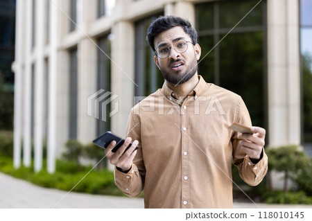 Man with glasses standing outside, holding phone and credit card, showing confused expression. Concept of financial uncertainty, technology frustration, decision making, or communication issues. Man with glasses standing outside, holding phone and credit card, showing confused expression. Concept of financial uncertainty, technology frustration, decision making, or communication issues. 118010145