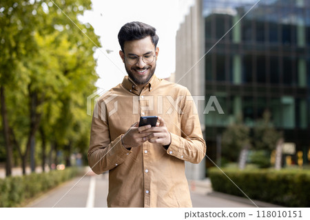 Smiling man wearing glasses and casual clothes using smartphone outdoors. Enjoying technology in urban setting with modern buildings. Capturing moment connectivity and communication through phone. 118010151