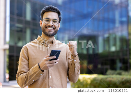 Smiling young man holding smartphone showing excitement and happiness in outdoor urban setting. Celebrating achievement or good news. Tech-savvy individual enjoying positive moment with phone Smiling young man holding smartphone showing excitement and happiness in outdoor urban setting. Celebrating achievement or good news. Tech-savvy individual enjoying positive moment with phone 118010351