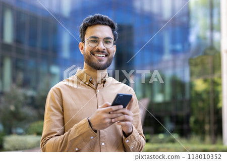 Confident young man with glasses holds smartphone while standing outside contemporary office. He is smiling at camera, suggesting positive communication and connectivity. 118010352