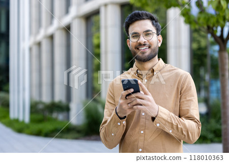 Smiling young man with glasses outside modern office building using smartphone. Dressed in casual outfit showcasing outdoor urban lifestyle. Concept of communication, technology, and modern business. 118010363