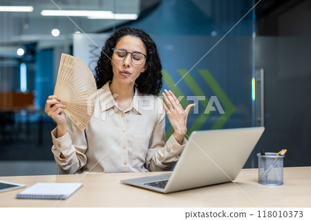 Hispanic businesswoman sits at office desk using laptop, cooling herself with hand fan. She expresses discomfort from heat, suggesting lack of air conditioning. Hispanic businesswoman sits at office desk using laptop, cooling herself with hand fan. She expresses discomfort from heat, suggesting lack of air conditioning. 118010373