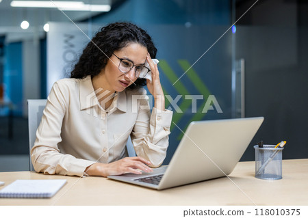 Hispanic businesswoman focused on laptop work, displaying concern in modern office setting. Professional attire and glasses emphasize dedication to task amidst working environment 118010375