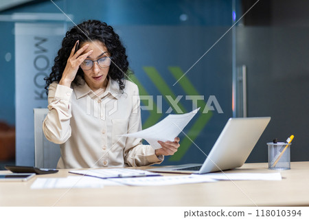 Stressed businesswoman analyzing document in office, seated at desk with laptop. Expression of worry. Concepts of work stress, problem solving, office environment. 118010394