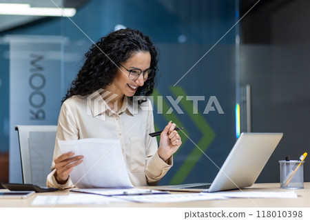Confident businesswoman with glasses working in office with laptop and documents. Phone and pen in hand convey productivity. Professional atmosphere emphasizes focus and dedication. 118010398