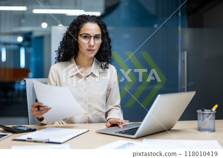 Confident businesswoman with glasses holding documents engaged in focused work at modern office desk. She uses laptop for financial analysis. Scene conveys professionalism, focus, and productivity 118010419