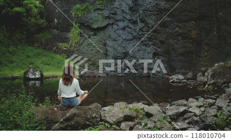 Woman practicing yoga near a small waterfall in a wild tropical jungle. She is enjoying a peaceful moment of mindfulness and connection with nature Woman practicing yoga near a small waterfall in a wild tropical jungle. She is enjoying a peaceful moment of mindfulness and connection with nature 118010704