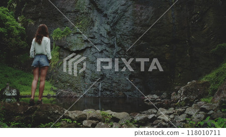 Young woman is enjoying the view of a waterfall cascading down a rocky cliff in a lush tropical forest. The serene atmosphere and the beauty of nature create a sense of peace and tranquility Young woman is enjoying the view of a waterfall cascading down a rocky cliff in a lush tropical forest. The serene atmosphere and the beauty of nature create a sense of peace and tranquility 118010711