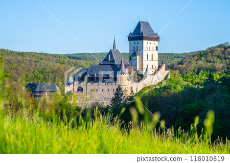 The majestic Karlstejn Castle stands proud against a backdrop of lush greenery as the morning sun rays enhance its architecture. A peaceful scene unfolds in this historic location of Czechia. 118010819