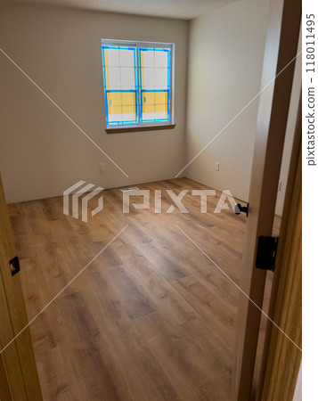 An empty room showcasing newly installed vinyl plank flooring. The space is under renovation, with windows covered in protective tape and neutral-colored walls. The view is from a hallway, through a An empty room showcasing newly installed vinyl plank flooring. The space is under renovation, with windows covered in protective tape and neutral-colored walls. The view is from a hallway, through a 118011495