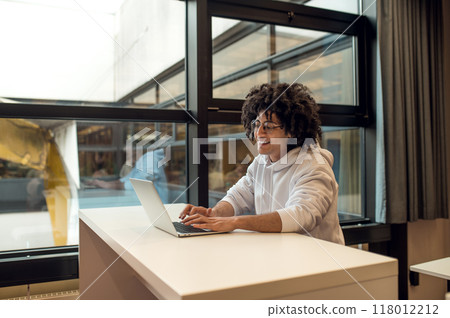 Young curly-haired man studying on his laptop in the college library 118012212