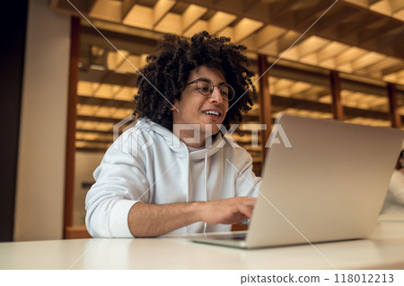 Male student watching webinar for his diploma project indoors via laptop in library Male student watching webinar for his diploma project indoors via laptop in library 118012213