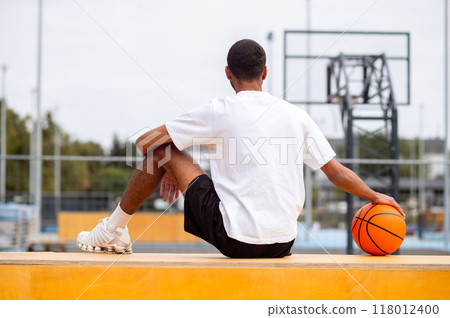 Man in white tshirt and with a ball sitting at the sports playground 118012400