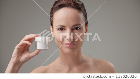 Beauty, health, cosmetics, anti-aging therapy and skin care concept - young beautiful brunette Caucasian woman holding a jar of moisturizer in her hands and looking at the camera 118020326