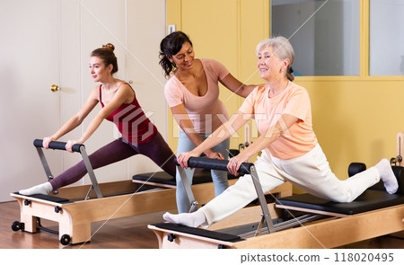 Aged woman doing pilates on reformer with Hispanic female trainer 118020495