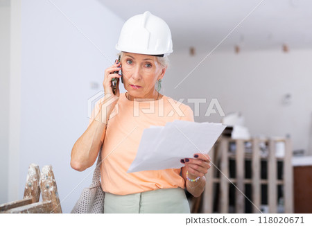 Elderly cottage owner in protective helmet talking on mobile phone in renovated room 118020671