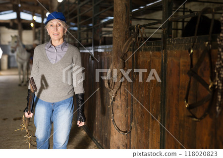 Smiling elderly woman carrying pitchfork to stack hay for horses in stable 118020823