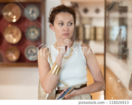 Woman with piece of paper and phone in hands walks around museum hall, view exhibition 118020913