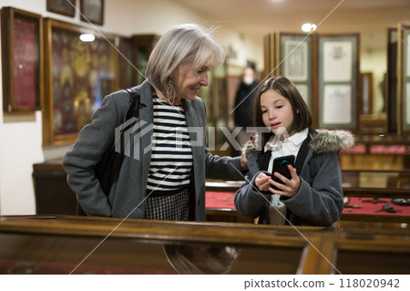 Tween girl with grandmother searching information about showpieces on smartphone in museum Tween girl with grandmother searching information about showpieces on smartphone in museum 118020942
