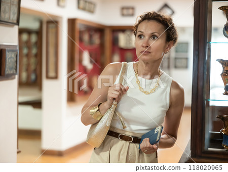 Woman with piece of paper and phone in hands walks around museum hall, view exhibition 118020965
