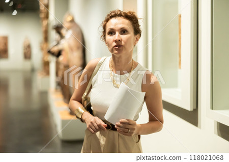 Interested young woman looking at collection of medieval sculptures and objects in museum 118021068