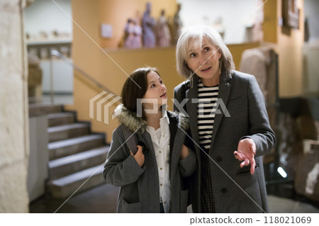 Aged female tutor with preteen girl exploring showpieces in museum 118021069
