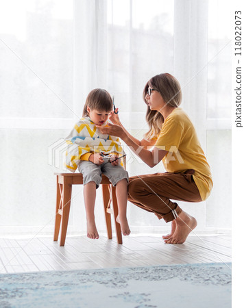 Mother cuts her son's hair by herself. Little boy sits, covered with cloth, and holds pair of scissors. Beauty at home lifestyle. 118022073