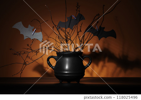 Mysterious black cauldron with dry plants and cutout bats in spotlight on pumpkin orange background. Represents Halloween, crafts, magic, and fantasy concepts. 118025496
