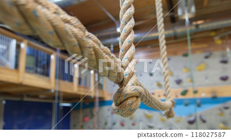 Close-up of a thick, knotted rope hanging indoors with a climbing wall and gym equipment in the background. 118027587