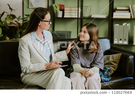 Counselor engaging with young girl on sofa in cozy office setting, fostering an environment of trust and empathy. Visible plants and book on shelf enhancing the warming ambiance 118027669