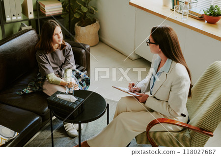 Counseling session within comfortable, cozy office setting with professional therapist taking notes. Client appears engaged in discussion, sitting on sofa beside indoor plants Counseling session within comfortable, cozy office setting with professional therapist taking notes. Client appears engaged in discussion, sitting on sofa beside indoor plants 118027687