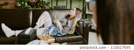 Woman reclining on sofa with closed eyes and relaxed posture. Cozy living room setting with plants, soft lighting, and comfortable furniture emphasizing peaceful atmosphere Woman reclining on sofa with closed eyes and relaxed posture. Cozy living room setting with plants, soft lighting, and comfortable furniture emphasizing peaceful atmosphere 118027695
