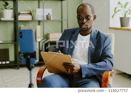 Portrait of businessman holding clipboard while sitting in modern office with shelves, plant and phone on tripod in background capturing video Portrait of businessman holding clipboard while sitting in modern office with shelves, plant and phone on tripod in background capturing video 118027757