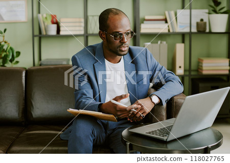 Man in blue suit and glasses conducting business meeting online while sitting on brown leather sofa in modern office with green walls and shelves behind Man in blue suit and glasses conducting business meeting online while sitting on brown leather sofa in modern office with green walls and shelves behind 118027765