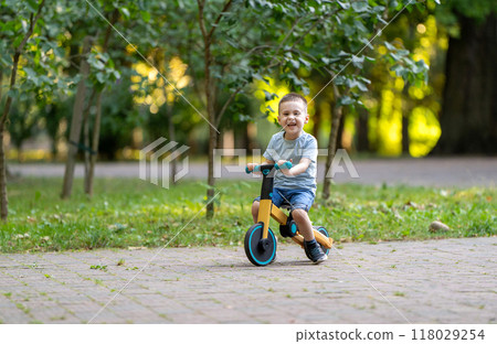A cute smiling toddler boy of three or four years old rides a bicycle or balance bike in a city park on a sunny summer day. Toddlerhood and childhood concept. Selective focus A cute smiling toddler boy of three or four years old rides a bicycle or balance bike in a city park on a sunny summer day. Toddlerhood and childhood concept. Selective focus 118029254