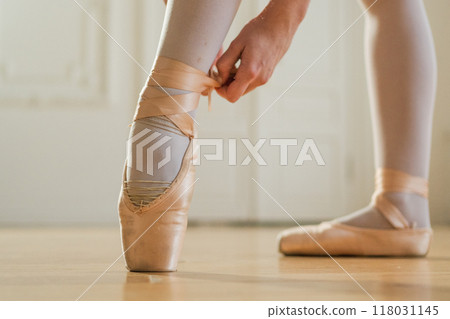 Ballerina puts on pointe shoes on leg in dance class. Classical ballet dancer woman in dance studio. Ballerina putting on tying classical pointe shoes for dance training. Ballet school concept 118031145