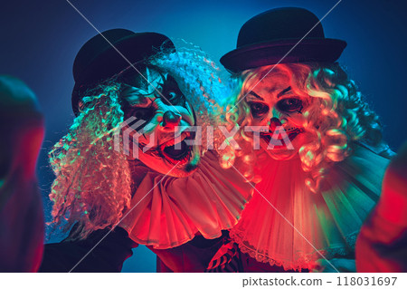 Portrait of creepy clown couple playfully posing looking at camera in neon light against gradient background. Portrait of creepy clown couple playfully posing looking at camera in neon light against gradient background. 118031697