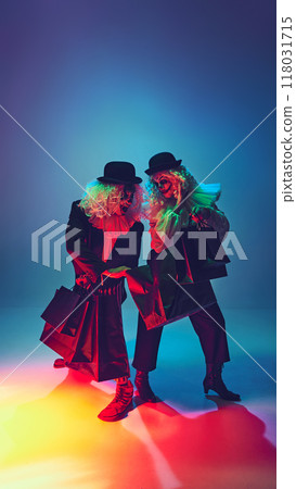 Clowns standing with shopping bags, their creepy smiles illuminated in neon light against gradient background. Furious shopaholics. Clowns standing with shopping bags, their creepy smiles illuminated in neon light against gradient background. Furious shopaholics. 118031715
