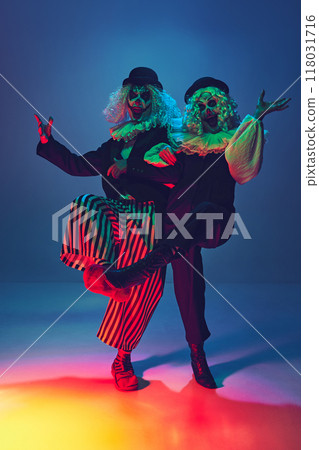 Crazy, frightening clowns with furious make-up dressed vintage attire dancing in neon light against gradient blue background. Crazy, frightening clowns with furious make-up dressed vintage attire dancing in neon light against gradient blue background. 118031716