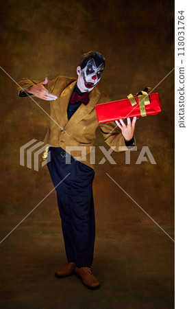 Proudly holding red gift, clown grins mischievously, as if presenting something dangerous against vintage dark backdrop. Proudly holding red gift, clown grins mischievously, as if presenting something dangerous against vintage dark backdrop. 118031764